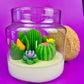 Cactus art candle in clear jar with cork lid; white wax with green cactus and blooms