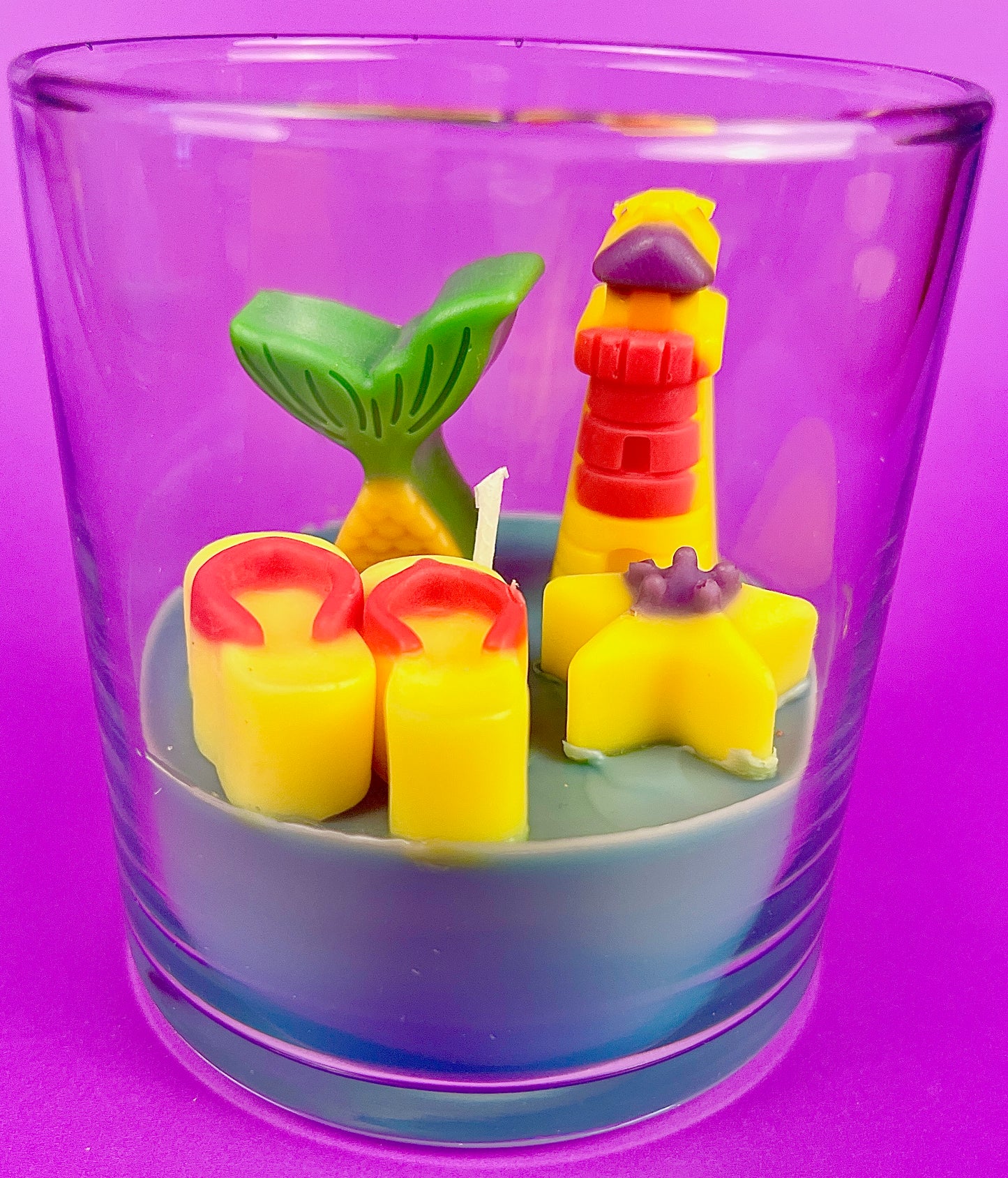 Small scented candle in clear glass jar with red-yellow lighthouse and yellow shell wax pieces