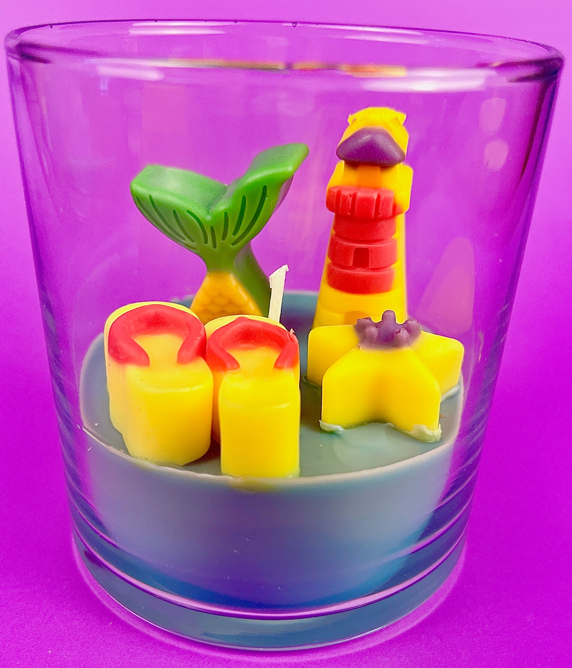 Small scented candle in clear glass jar with red-yellow lighthouse and yellow shell wax pieces