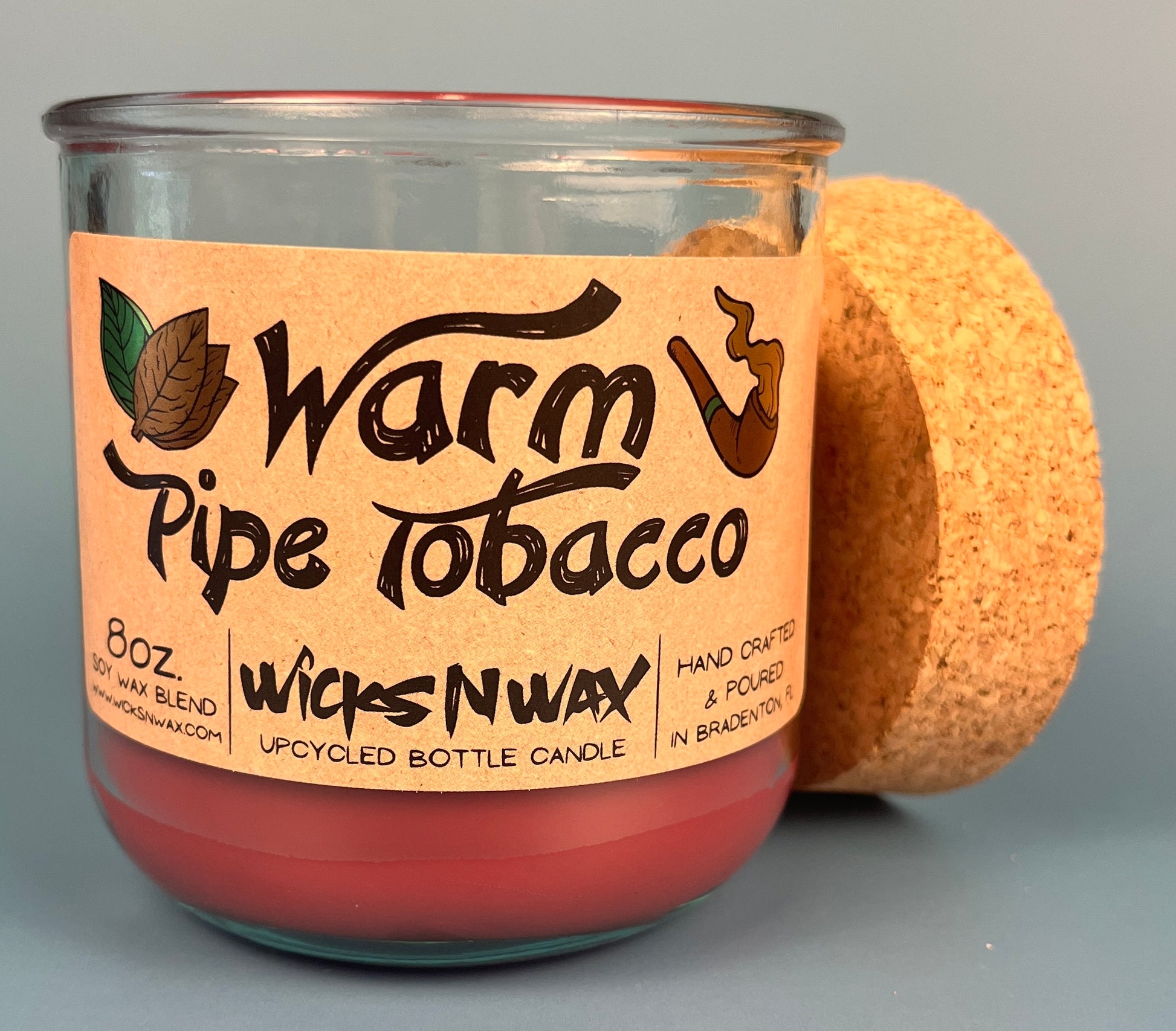Glass bottle candle with red wax, leaf-and-smoke graphic label, cork lid behind