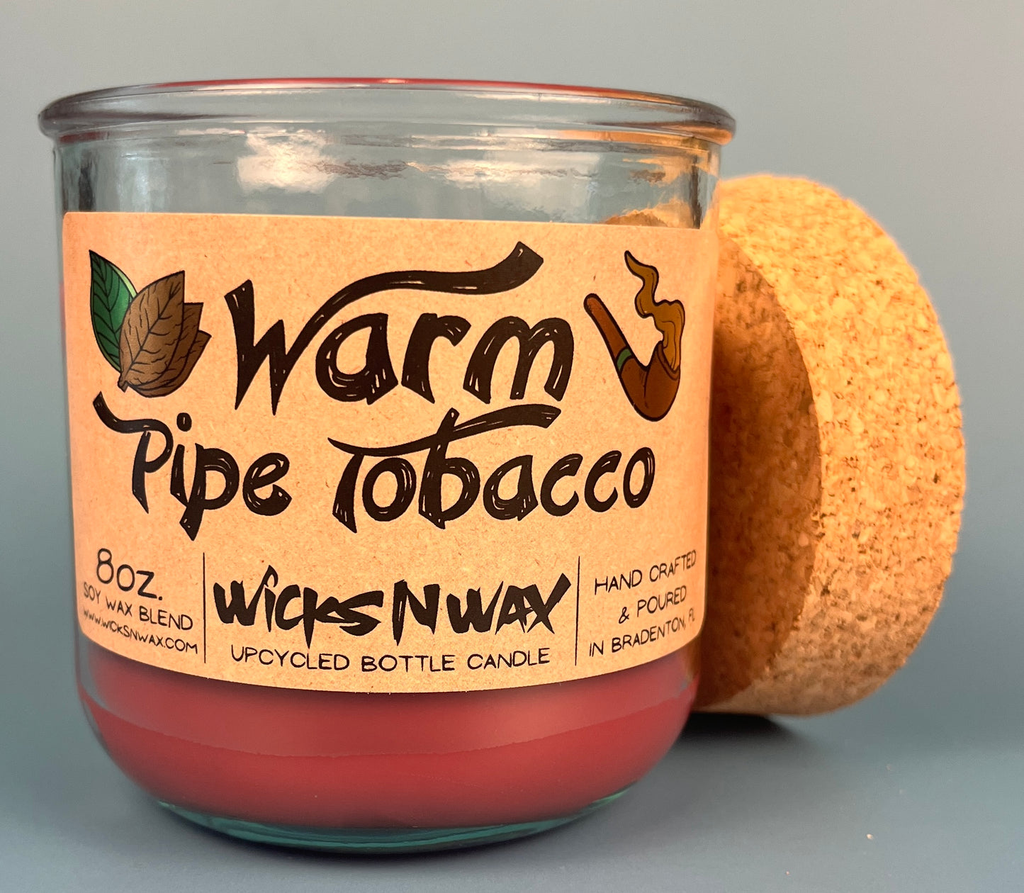 Glass bottle candle with red wax and leaf-and-smoke graphic label, cork lid behind