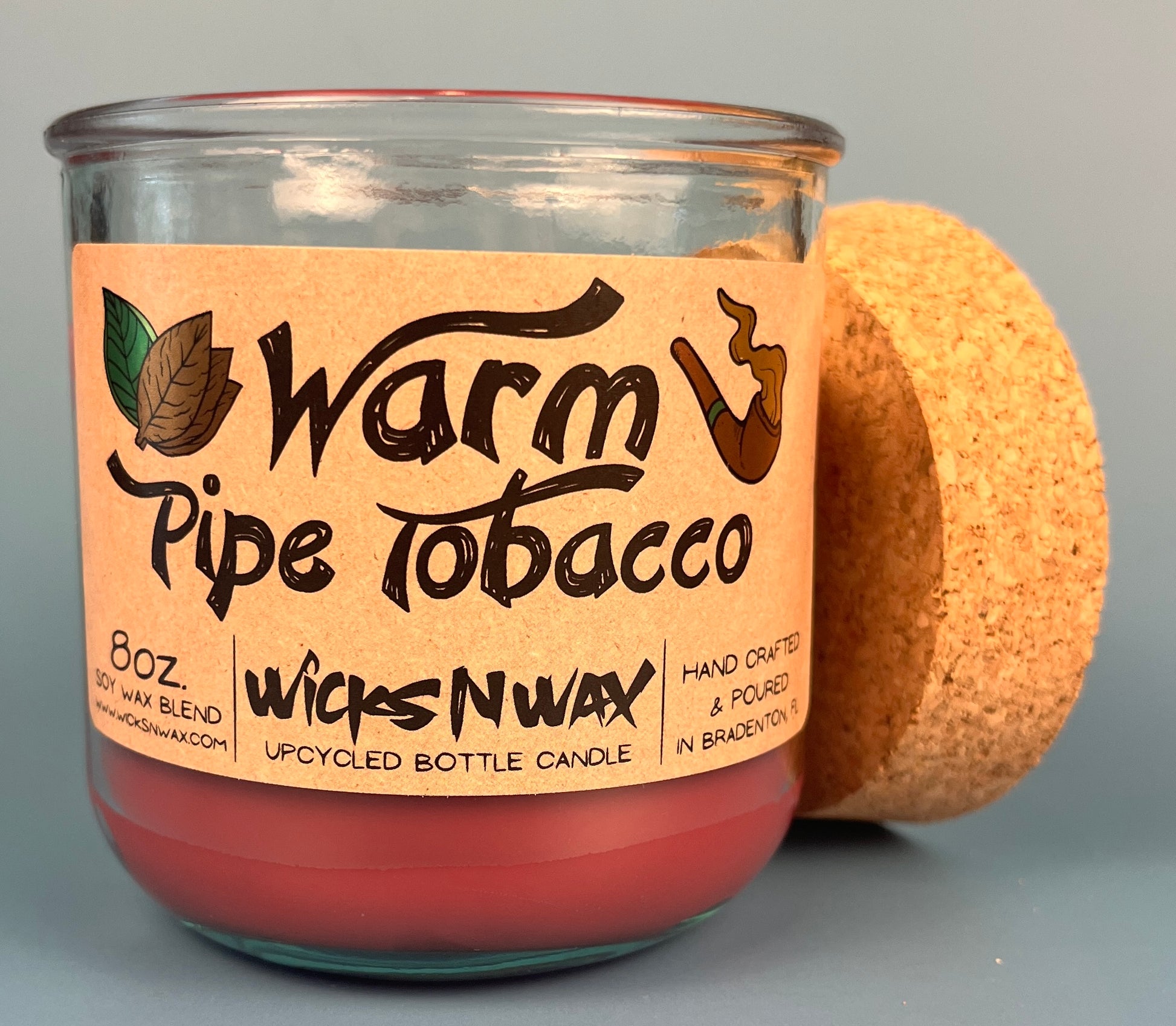 Glass bottle candle with red wax and leaf-and-smoke graphic label, cork lid behind